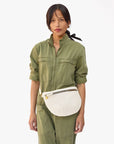 A person stands against a plain background, wearing an olive green jumpsuit with a high collar and front pockets. They carry the Clare Vivier Grande Fanny Perf, a white crossbody bag with an adjustable strap and zipper. Their hair is tied back with a black ribbon.