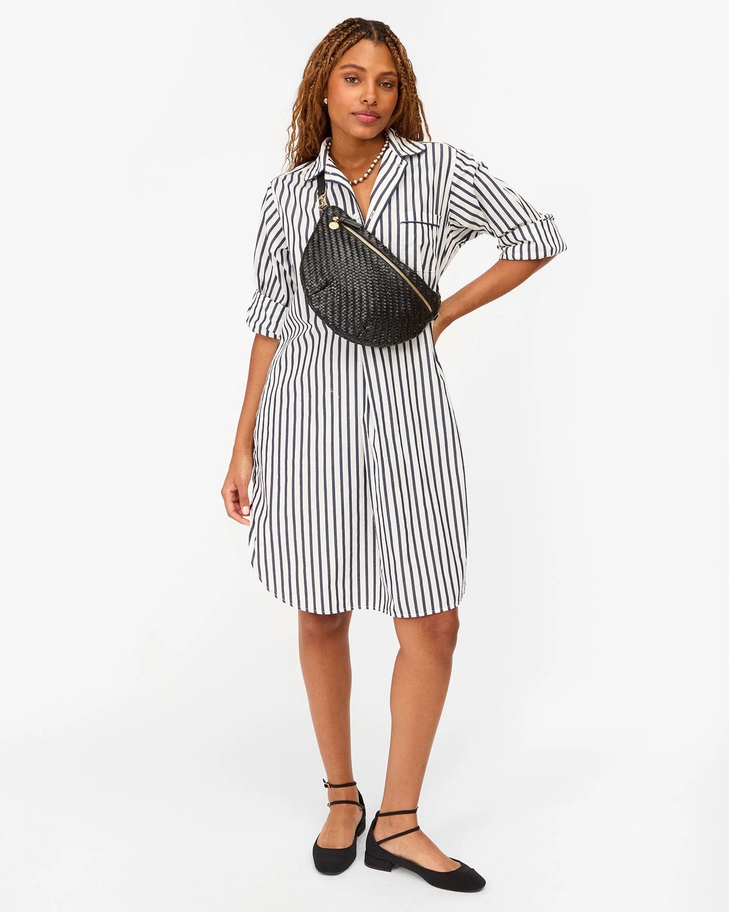 A woman with long, wavy hair poses against a white background, wearing a white shirt dress with black stripes, black flats, and the Grande Fanny S25 by Clare Vivier—a handwoven leather crossbody bag with an adjustable strap.