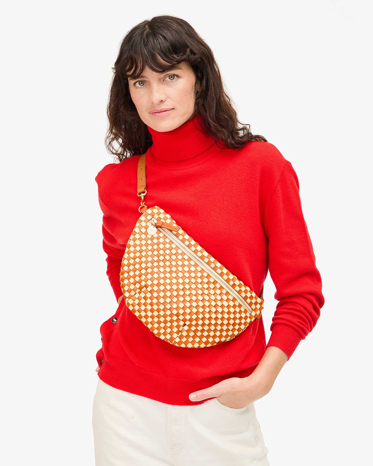 A person with long dark hair, dressed in a red turtleneck and white pants, effortlessly holds the adjustable strap of Clare Vivier&#39;s Grande Fanny Woven Checker crossbody bag against a plain white background.