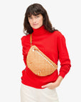 A person with long dark hair, dressed in a red turtleneck and white pants, effortlessly holds the adjustable strap of Clare Vivier's Grande Fanny Woven Checker crossbody bag against a plain white background.