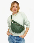 A person with short hair, wearing a light green sweater and denim jeans, poses with the Grande Fanny Zig-Zag bag from Clare Vivier. They have a relaxed expression with their hands in their pockets against a plain white background.