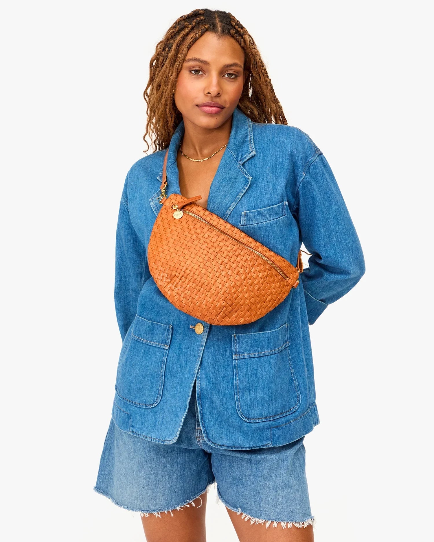 A woman with long braids wears a blue denim jacket and shorts against a white background, carrying the Clare Vivier Grande Fanny S25—a large handwoven leather crossbody bag with adjustable strap—slung across her chest.