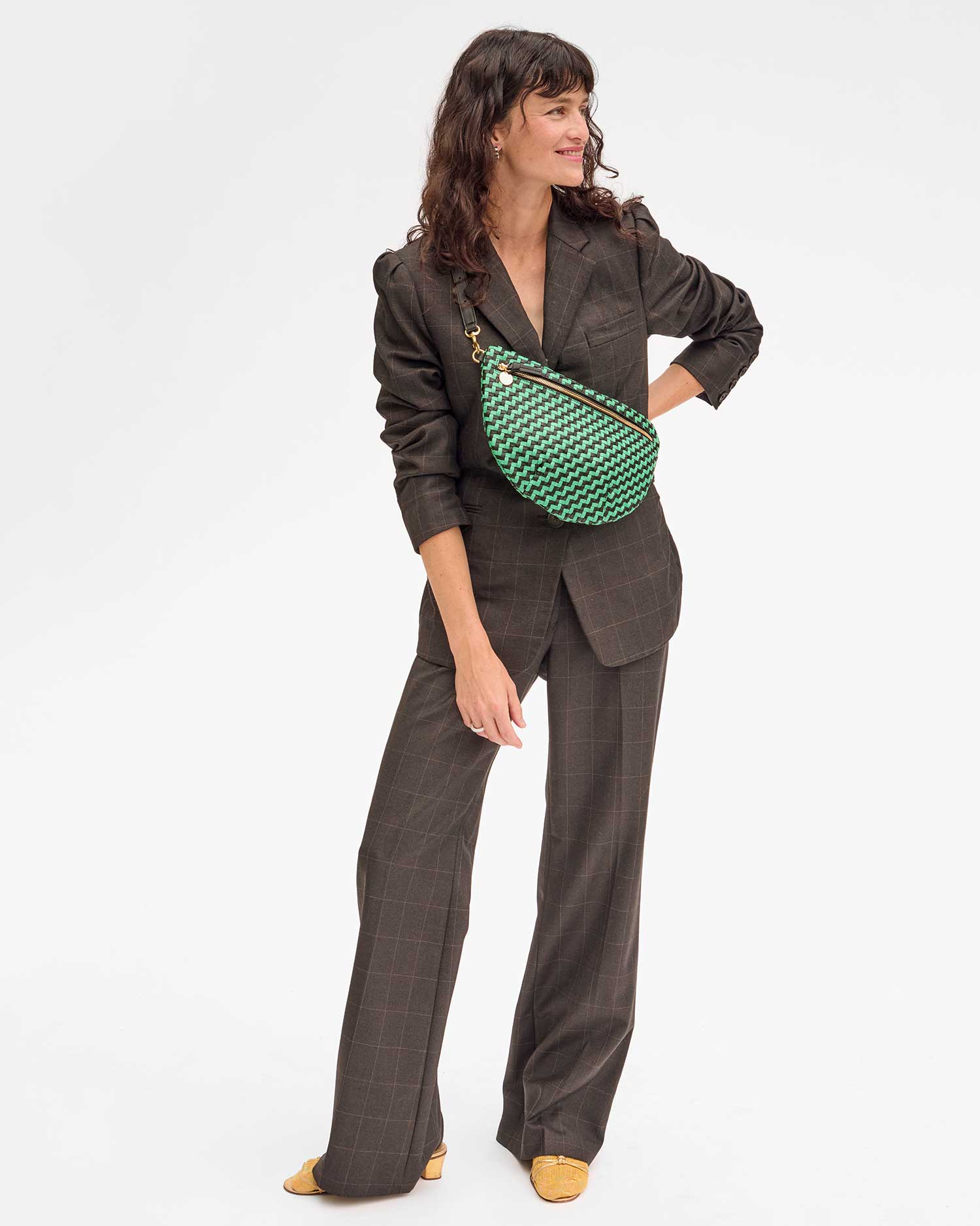 A woman in a brown plaid suit stands smiling, carrying the stylish Grande Fanny Zig-Zag crossbody bag by Clare Vivier, featuring an adjustable strap and a green and black checkered pattern. She has wavy hair and wears yellow shoes, all set against a plain white background.