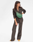 A woman in a brown plaid suit stands smiling, carrying the stylish Grande Fanny Zig-Zag crossbody bag by Clare Vivier, featuring an adjustable strap and a green and black checkered pattern. She has wavy hair and wears yellow shoes, all set against a plain white background.