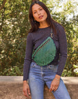 A person with long dark hair leans against a stone ledge outdoors, wearing a black long-sleeve top and blue jeans. Positioned stylishly at their side is the Grande Fanny Zig-Zag crossbody bag by Clare Vivier, featuring an adjustable strap and green zigzag pattern. In the background, lush green foliage flourishes.