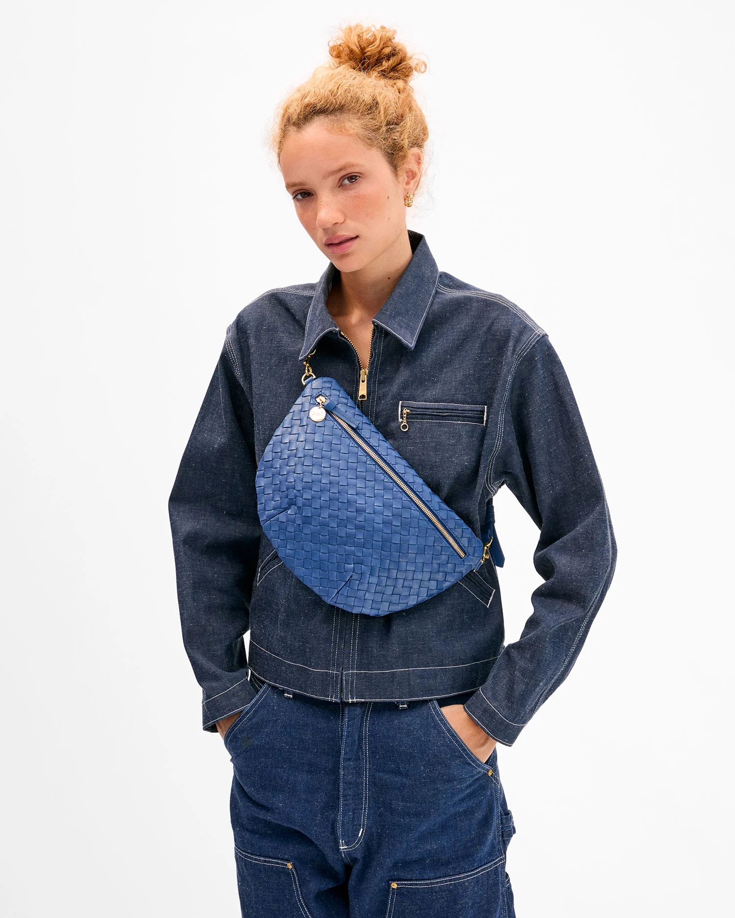 A woman with curly hair in a bun, wearing a dark denim jacket and pants, carries the Clare Vivier Grande Fanny S26—an oversized handwoven leather crossbody bag—across her chest against a plain white background.