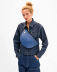 A woman with curly hair in a bun, wearing a dark denim jacket and pants, carries the Clare Vivier Grande Fanny S26—an oversized handwoven leather crossbody bag—across her chest against a plain white background.