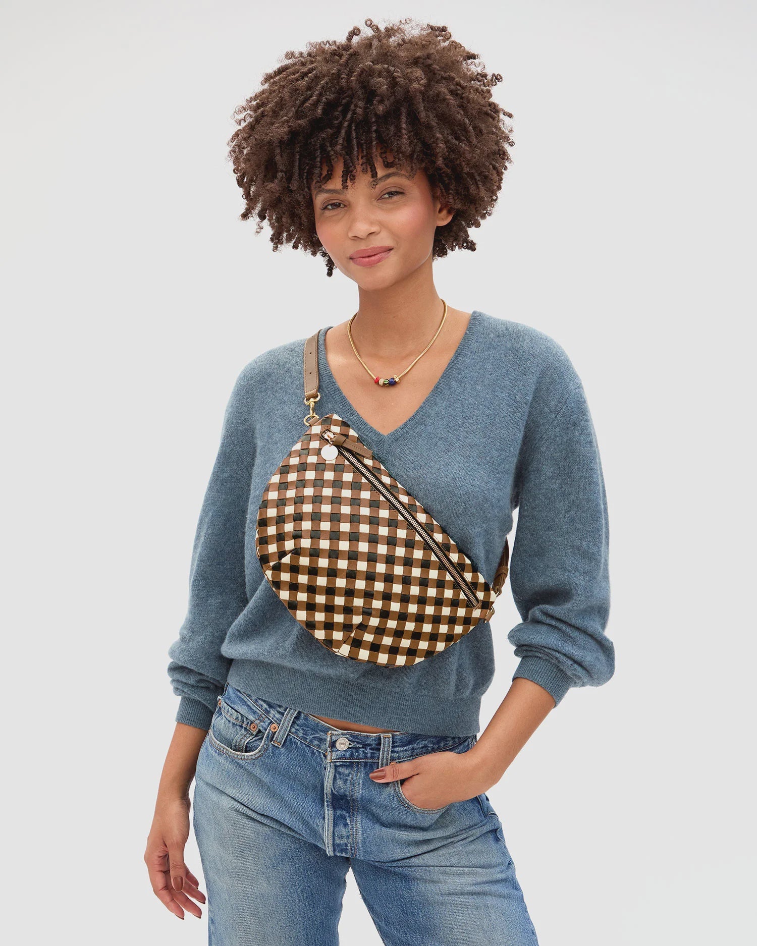 A person in a blue V-neck sweater and jeans models the Clare Vivier Grande Fanny S26, a brown and white checkered handwoven leather crossbody bag, with a beaded necklace against a plain white background.