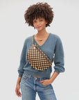 A person in a blue V-neck sweater and jeans models the Clare Vivier Grande Fanny S26, a brown and white checkered handwoven leather crossbody bag, with a beaded necklace against a plain white background.