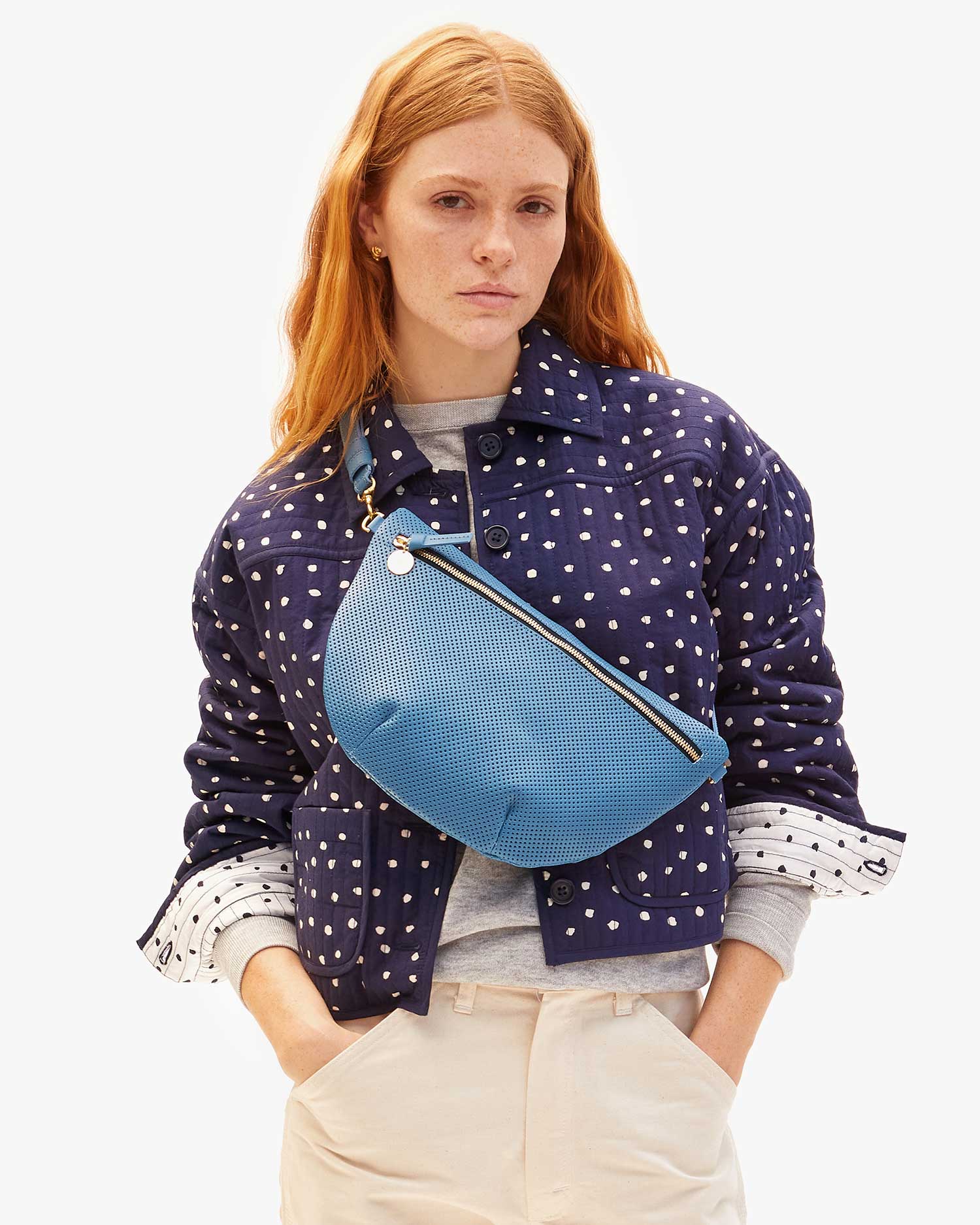 A person with long red hair wears a blue polka-dotted jacket over a gray shirt and beige pants, carrying Clare Vivier's stylish Grande Fanny Perf, featuring an adjustable strap and textured surface, set against a plain white backdrop.