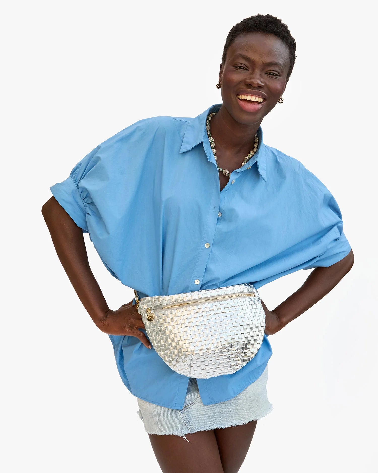 A smiling woman stands with her hands on her hips against a white background, wearing a loose blue button-up shirt, light denim shorts, a chunky necklace, and the Clare Vivier Grande Fanny S25 handwoven leather bag with adjustable strap.