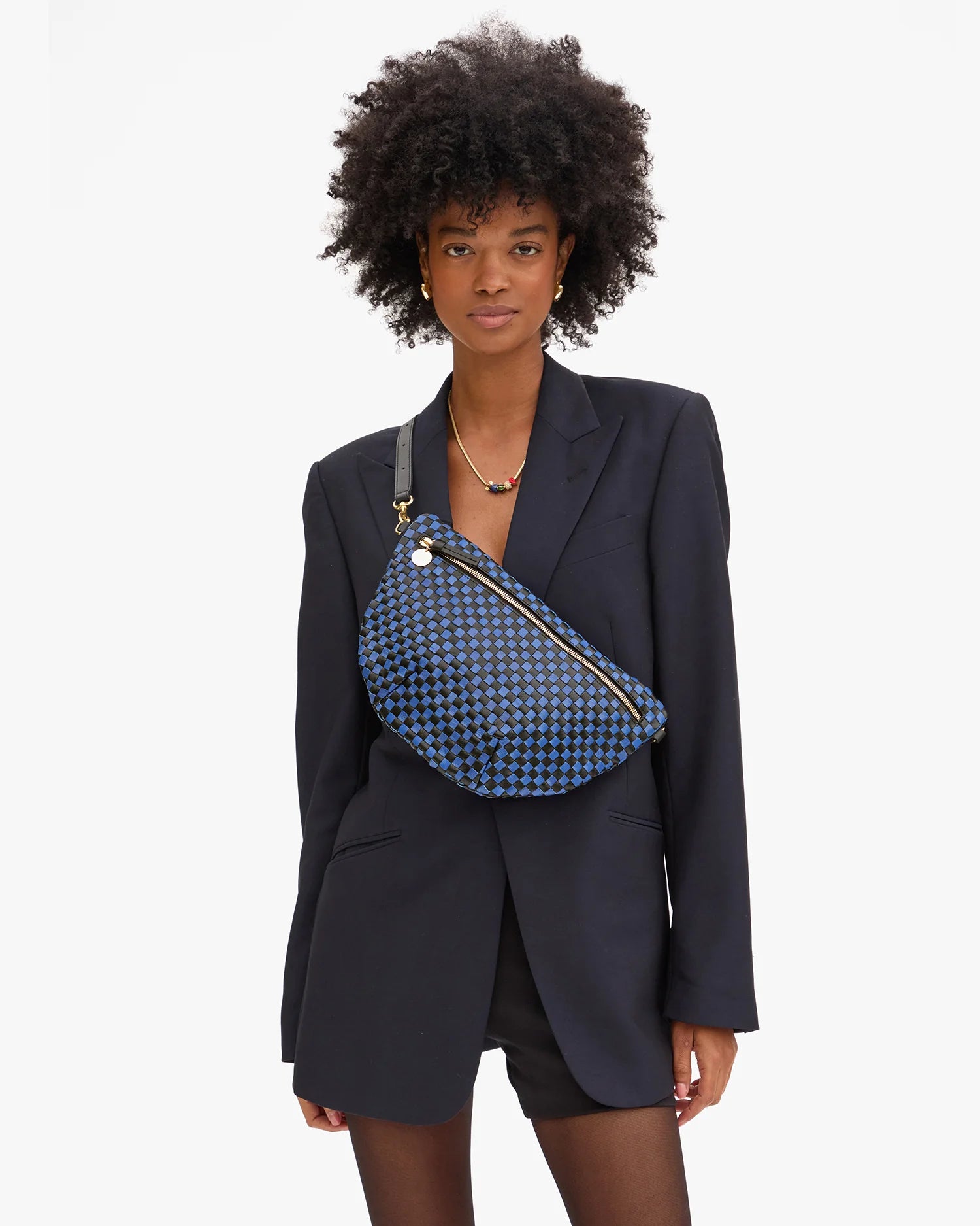 A woman in a black blazer and shorts stands against a plain background, wearing her natural curly hair and carrying the Clare Vivier Grande Fanny S25, a blue patterned crossbody bag with an adjustable strap.
