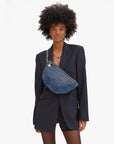 A woman in a black blazer and shorts stands against a plain background, wearing her natural curly hair and carrying the Clare Vivier Grande Fanny S25, a blue patterned crossbody bag with an adjustable strap.