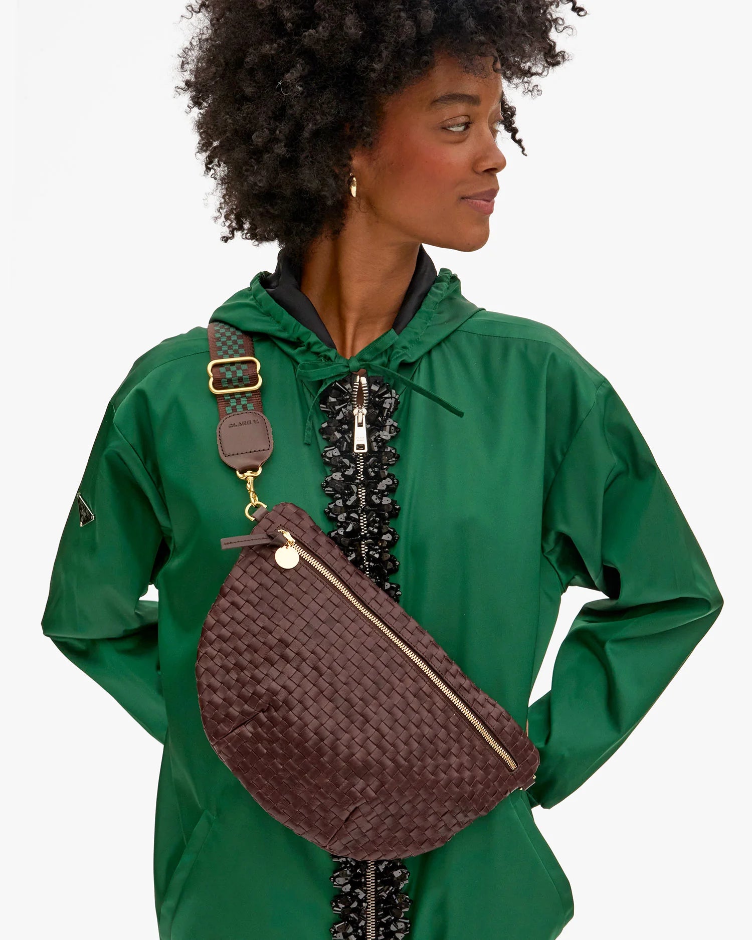 A woman wears a green jacket and a large dark brown Clare Vivier crossbody bag with gold hardware and an Adjustable Crossbody Strap, looking right with hands on hips.