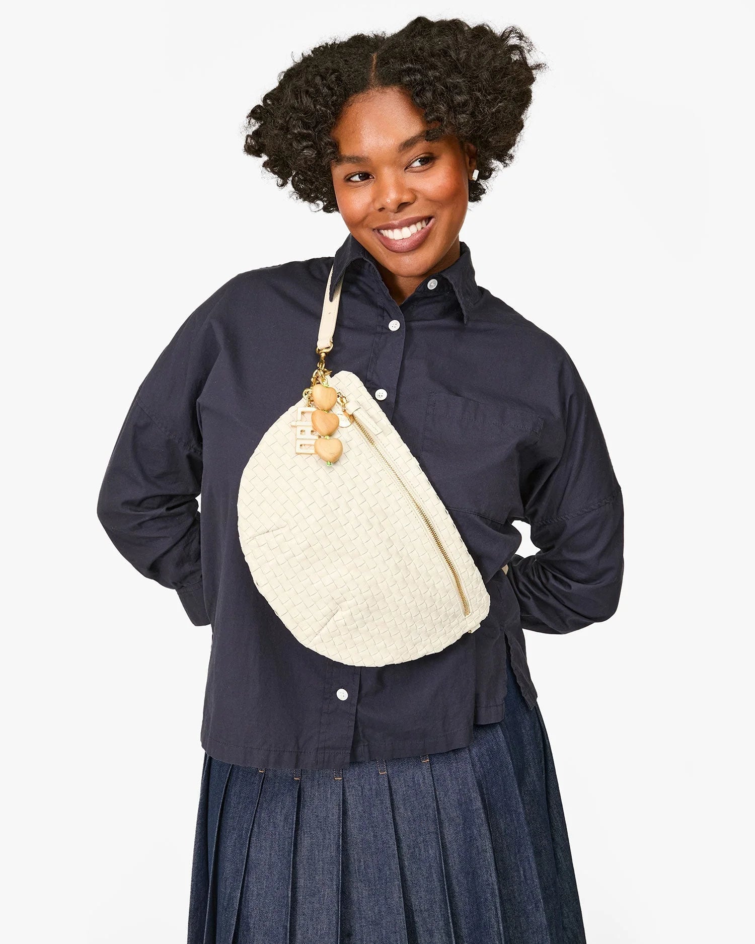 A smiling person with curly hair wears a navy button-up shirt, a pleated skirt, and carries the Clare Vivier Grande Fanny S25, a handwoven leather crossbody bag with an adjustable strap and wooden charms.