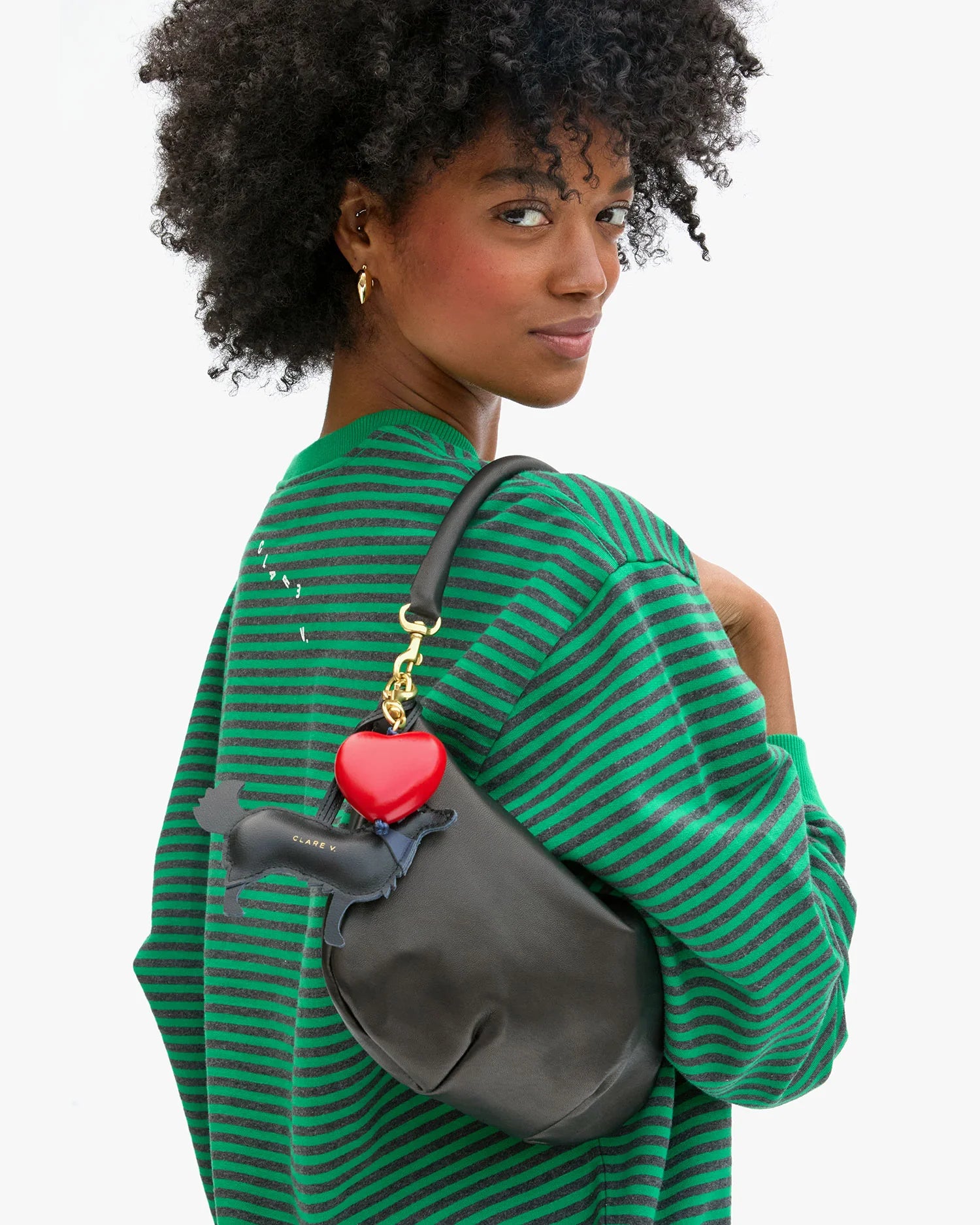 A woman with curly hair in a green and black striped top carries a smooth leather handbag from Clare Vivier, adorned with a red heart keychain, a small wallet, and the Coqui Fob accessory.