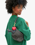 A woman with curly hair in a green and black striped top carries a smooth leather handbag from Clare Vivier, adorned with a red heart keychain, a small wallet, and the Coqui Fob accessory.