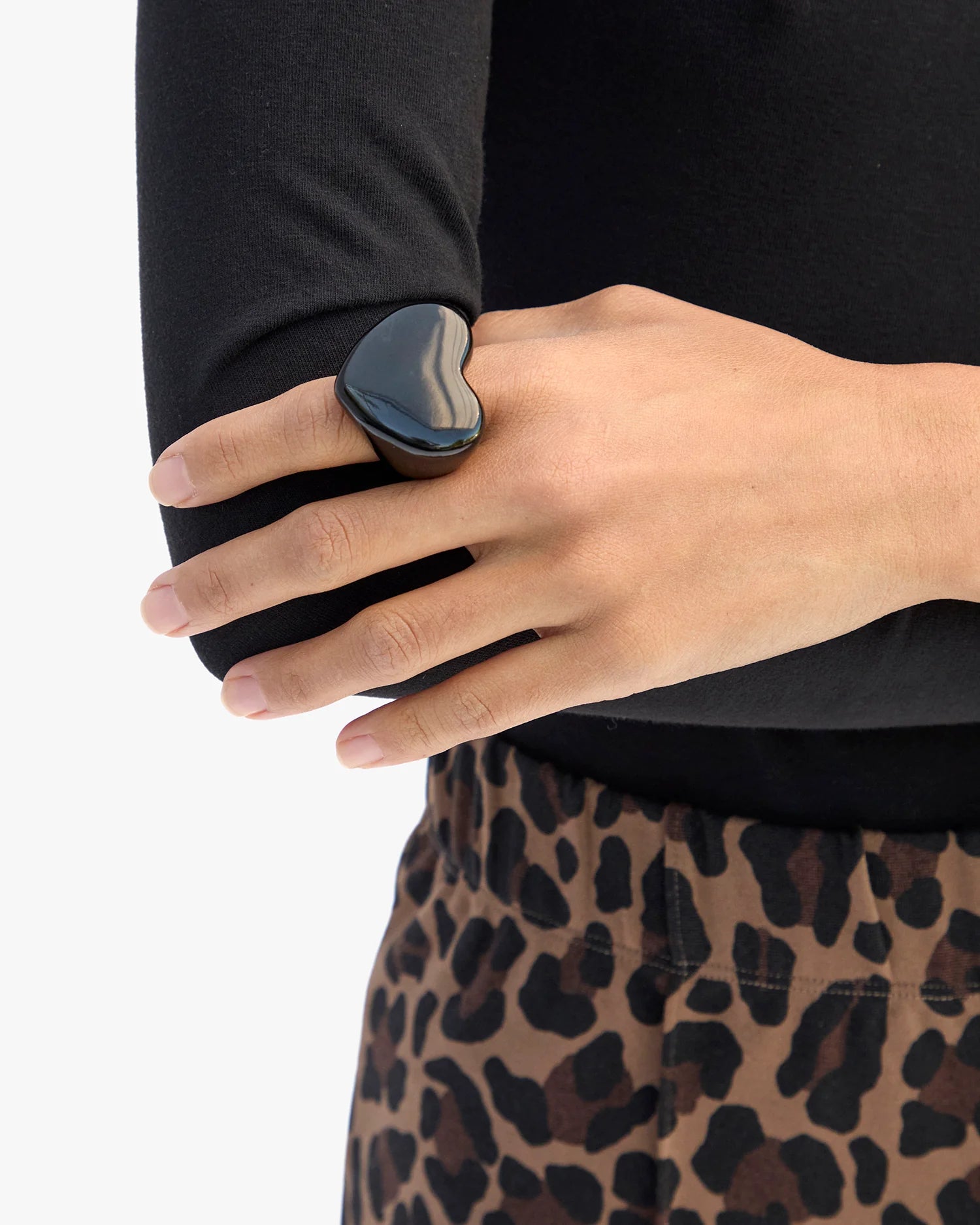 A person in leopard-print pants and a black top rests their left hand on their right arm, showcasing the Grande Heart Ring / Black by Clare Vivier (size 7) on their index finger—a chic accessory that completes the look.