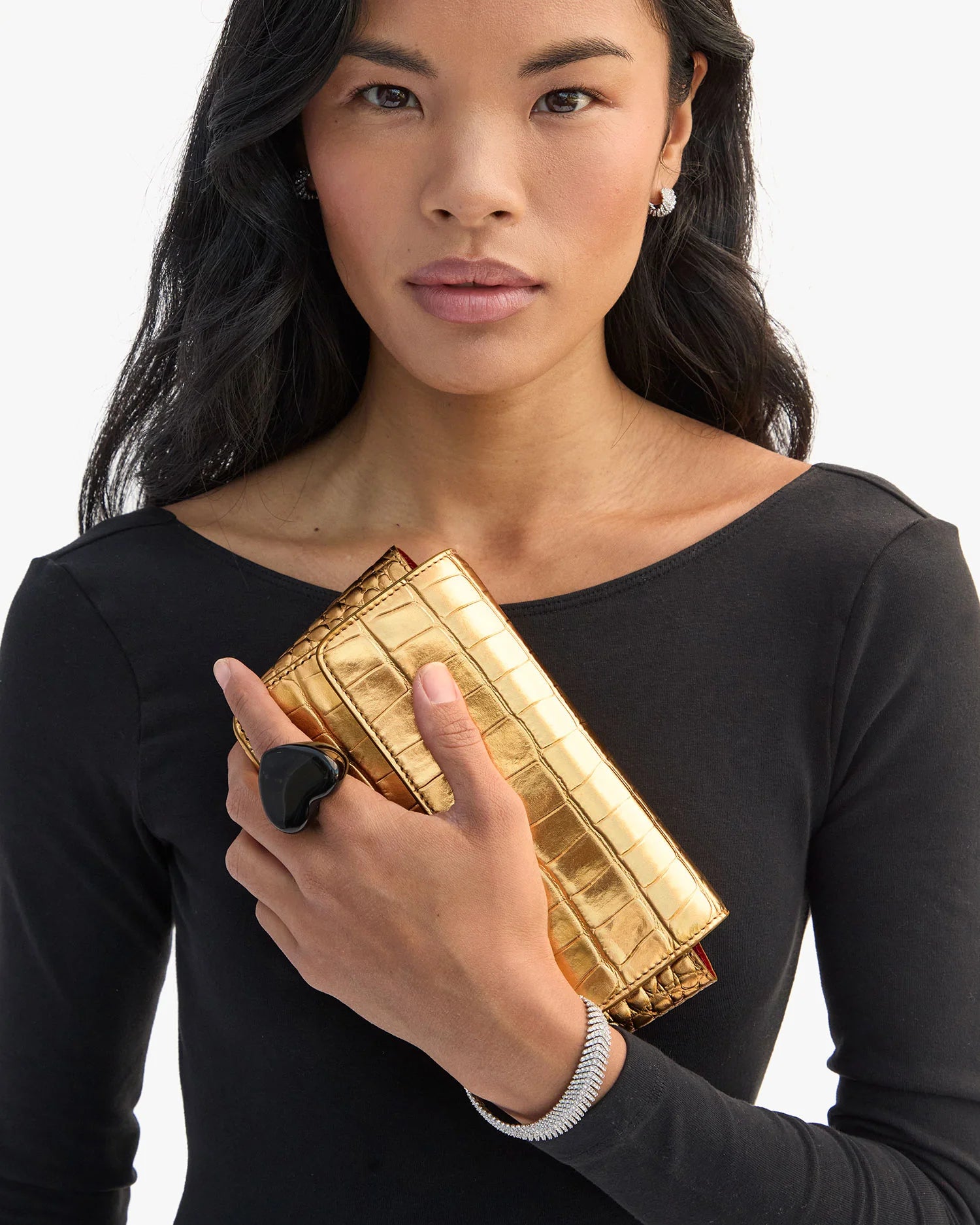 A woman with long black hair wears a black top, holds a shiny gold crocodile-patterned clutch, and models Clare Vivier’s Grande Heart Ring / Black (size 7) with a silver bracelet. She gazes at the camera against a white background.