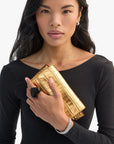 A woman with long black hair wears a black top, holds a shiny gold crocodile-patterned clutch, and models Clare Vivier’s Grande Heart Ring / Black (size 7) with a silver bracelet. She gazes at the camera against a white background.