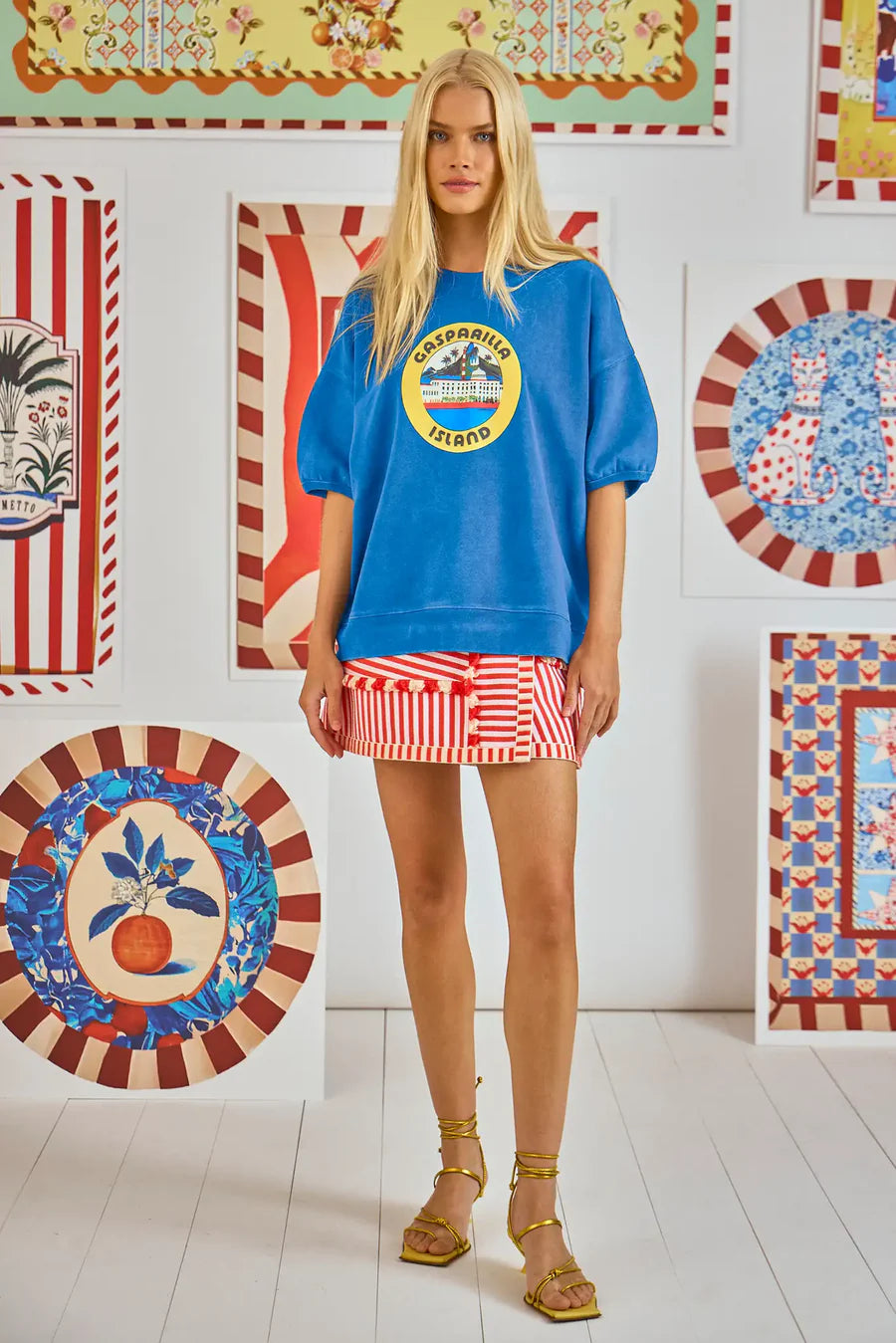 In a vibrant room with framed artworks, a long-haired blonde woman wears a Hunter Bell NYC Graphic Sweatshirt made of cozy French terry, paired with a red-and-white striped mini skirt and yellow strappy sandals.