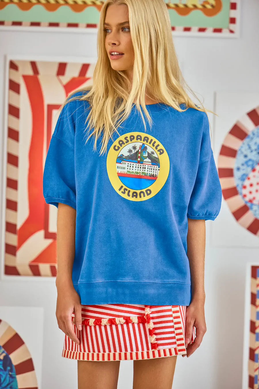 A woman with long blonde hair is wearing a blue oversized "Hunter Bell NYC" Graphic Sweatshirt and a red-and-white striped skirt, standing against a colorful abstract background that offers a vintage-inspired vibe.