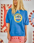 A woman with long blonde hair is wearing a blue oversized "Hunter Bell NYC" Graphic Sweatshirt and a red-and-white striped skirt, standing against a colorful abstract background that offers a vintage-inspired vibe.