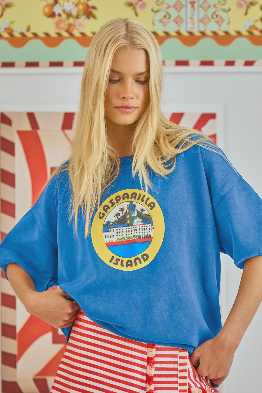 A blonde woman in an oversized Hunter Bell NYC Graphic Sweatshirt with "Gasparilla Island" text and red and white striped pants stands before a vibrant, floral-patterned background.