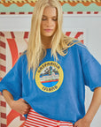 A blonde woman in an oversized Hunter Bell NYC Graphic Sweatshirt with "Gasparilla Island" text and red and white striped pants stands before a vibrant, floral-patterned background.