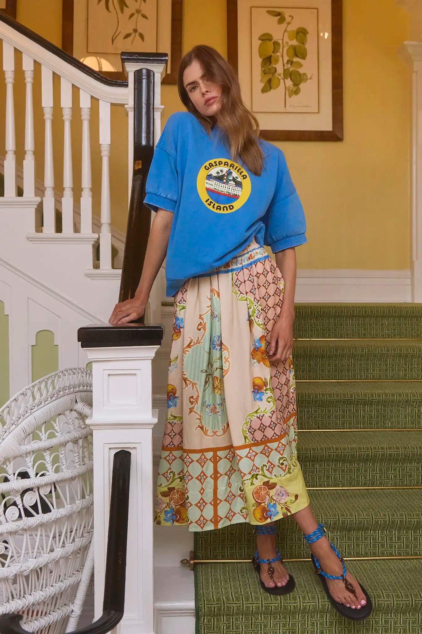 A person leans on a staircase railing, wearing a blue T-shirt with a yellow circular logo and Hunter Bell NYC's vibrant Tatum Skirt in Gasparilla Gardens print. Strappy sandals adorn their feet. The staircase is covered in green carpeting, with framed botanical prints on the wall.