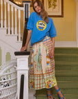 A person leans on a staircase railing, wearing a blue T-shirt with a yellow circular logo and Hunter Bell NYC's vibrant Tatum Skirt in Gasparilla Gardens print. Strappy sandals adorn their feet. The staircase is covered in green carpeting, with framed botanical prints on the wall.