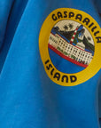 A close-up of Hunter Bell NYC's Graphic Sweatshirt shows a blue, vintage-inspired design. It features "Gasparilla Island" text encircling a coastal scene of a lighthouse, palm trees, and buildings set within a yellow border.