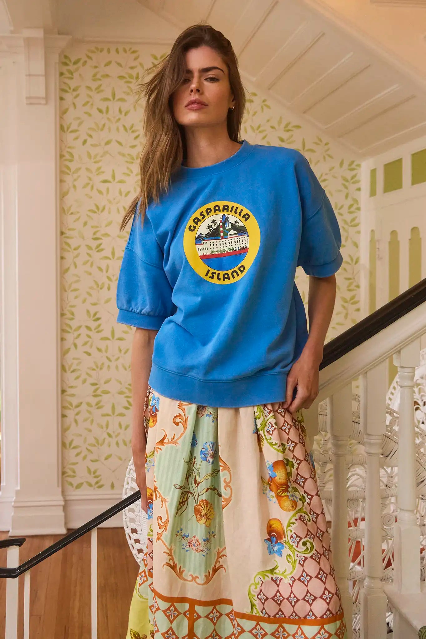 A woman poses on a staircase, wearing Hunter Bell NYC's blue Graphic Sweatshirt crafted from cozy French Terry fabric, paired with a vibrant skirt. Her long hair cascades gracefully as she stands against floral patterned wallpaper, with one hand confidently on her hip.