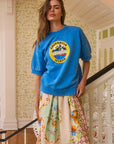 A woman poses on a staircase, wearing Hunter Bell NYC's blue Graphic Sweatshirt crafted from cozy French Terry fabric, paired with a vibrant skirt. Her long hair cascades gracefully as she stands against floral patterned wallpaper, with one hand confidently on her hip.