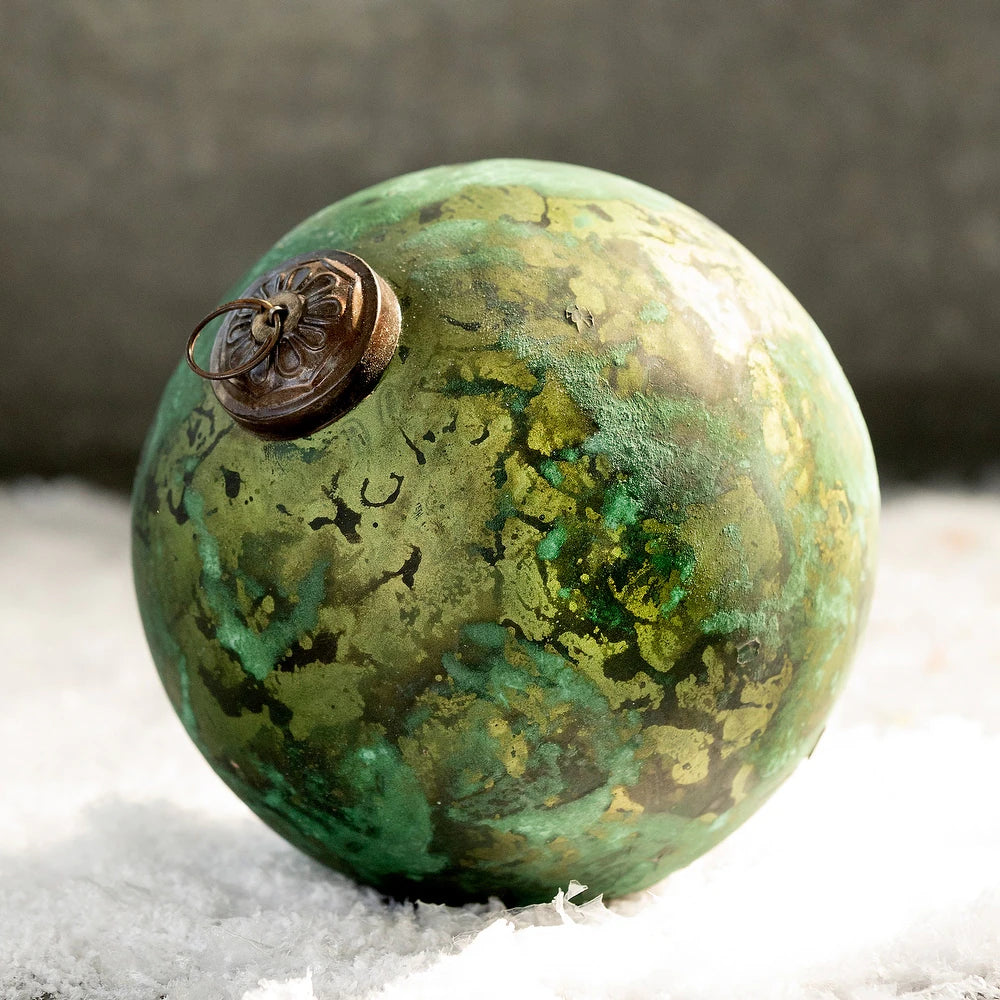 The Creative Co-op Marbled Glass Ornament showcases a green, patina-inspired finish with dark green and brown streaks and a metallic cap, resting elegantly on snowy surfaces.