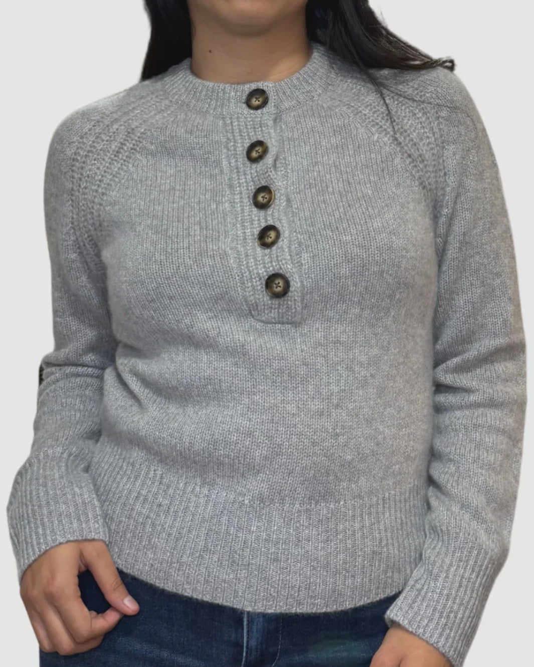 A person wears the White + Warren Cashmere Luxe Henley in light gray, featuring five brown buttons diagonally from collarbone to chest, paired with dark blue jeans against a plain white background.