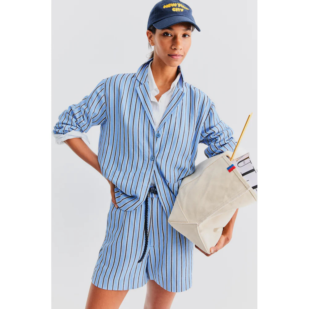 Wearing The Gwen by Kule, with a pajama-inspired stripe shirt and shorts, they roll up their sleeves for a casual flair. A navy cap completes the look while a beige tote brimming with essentials finalizes the ensemble against a plain white backdrop.