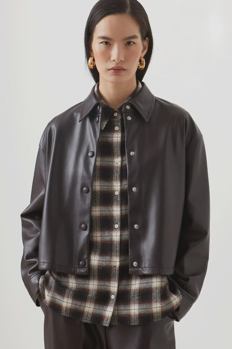 A woman with straight dark hair wears the SUNCOO LENNOX black pleather jacket over a black and beige plaid button-up shirt, paired with dark trousers and gold hoop earrings, standing against a plain light background.