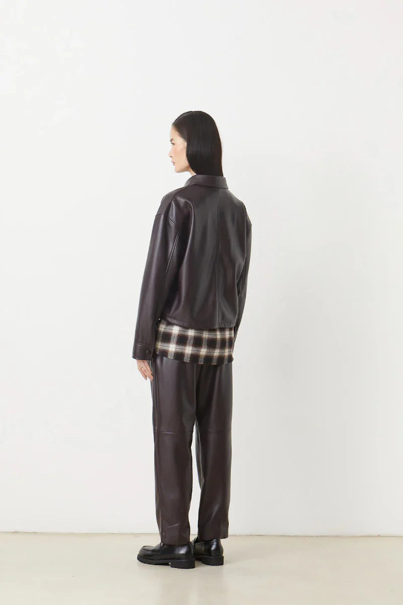 A person with straight, dark hair faces away, wearing the LENNOX Jacket by SUNCOO in dark brown pleather and matching pants. A plaid shirt peeks out beneath the jacket against a plain white background.