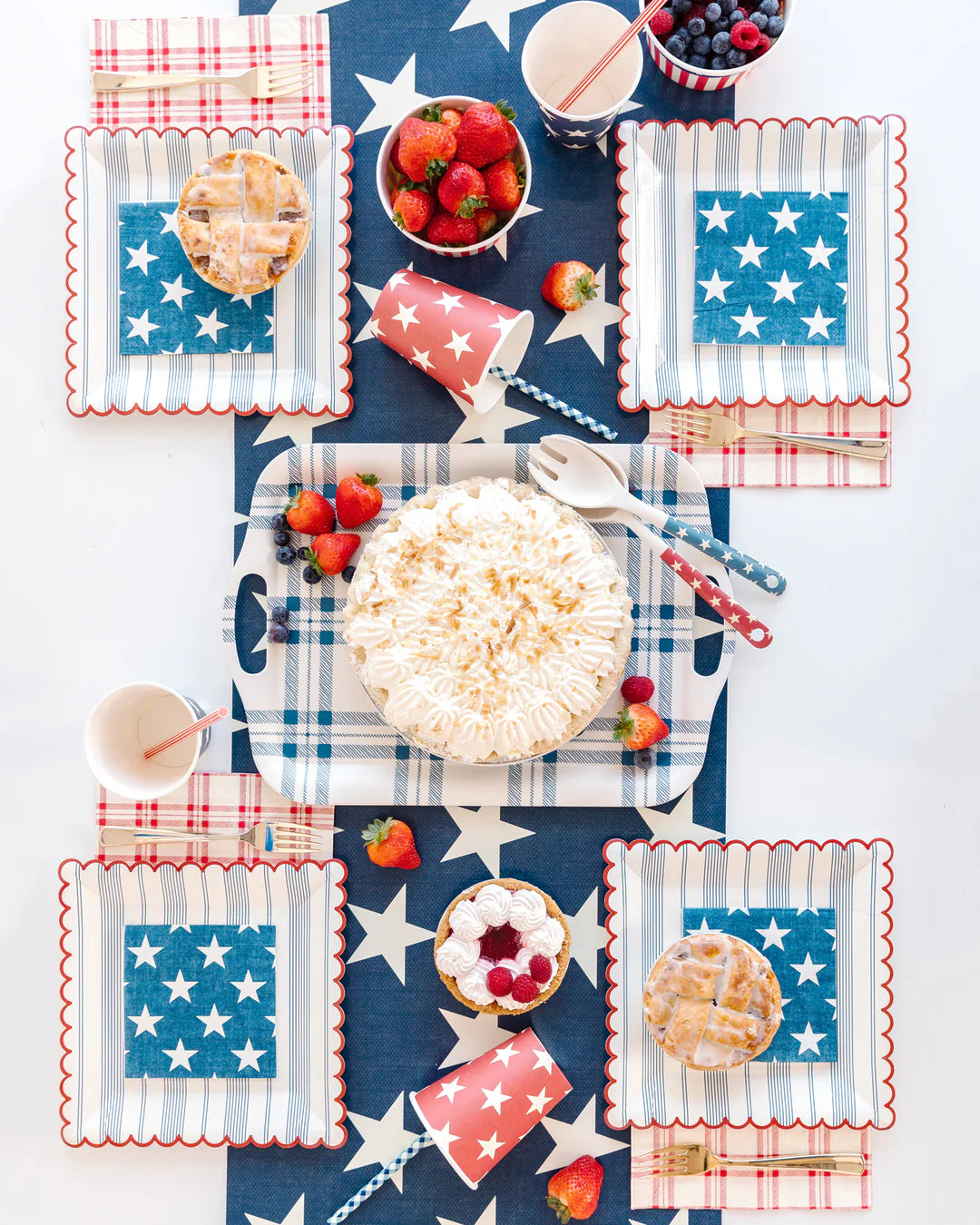 A festive summer barbeque table with My Mind's Eye Cocktail Paper Napkins, star-spangled plates, a whipped cream pie, mini pies, berries, pink drinks, and red, white & blue decor for a holiday celebration.