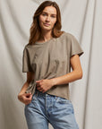 A person with long hair is wearing the Harley SS Boxy Crew in taupe from Perfectwhitetee, alongside mid-rise denim light blue jeans. They stand against a neutral draped curtain, looking directly at the camera.