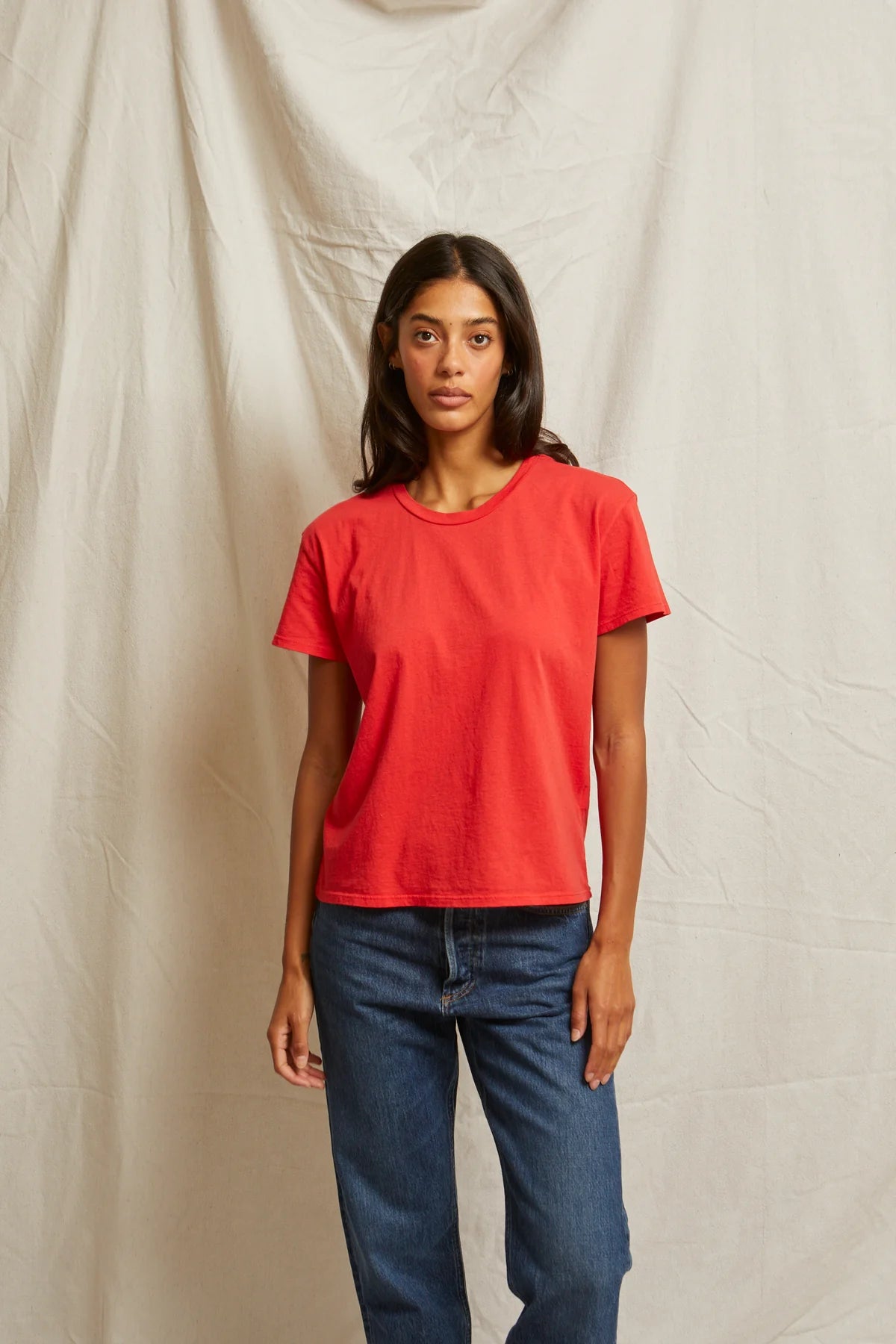 A person with long dark hair stands against a beige fabric backdrop, wearing the Perfectwhitetee Harley SS Boxy Crew in red and mid-rise denim jeans, looking at the camera with a neutral expression.