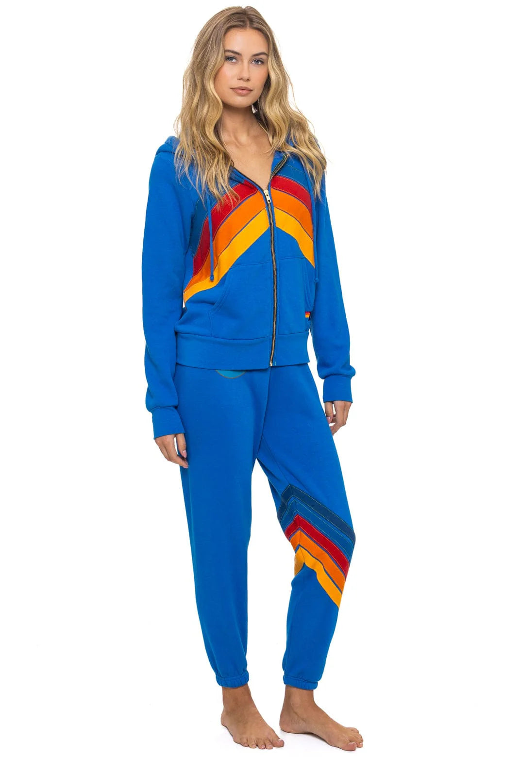 A woman with long, wavy blonde hair and a neutral expression stands barefoot wearing Aviator Nation's all seasons circle chevron women's sweatpant, styled in a bright blue with vintage-inspired chevron stripes.