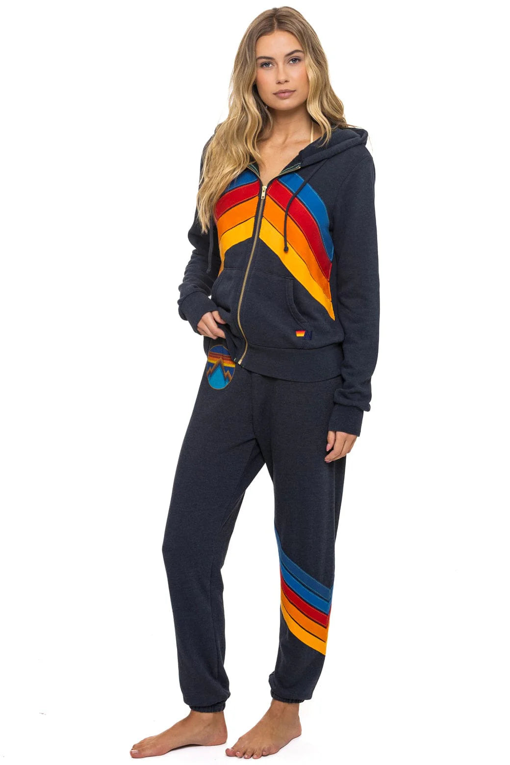 A woman with long blonde hair and a neutral expression stands barefoot in Aviator Nation’s all seasons circle chevron women’s sweatpants, featuring bold diagonal stripes for a vintage vibe, styled casually with a matching zip-up hoodie.