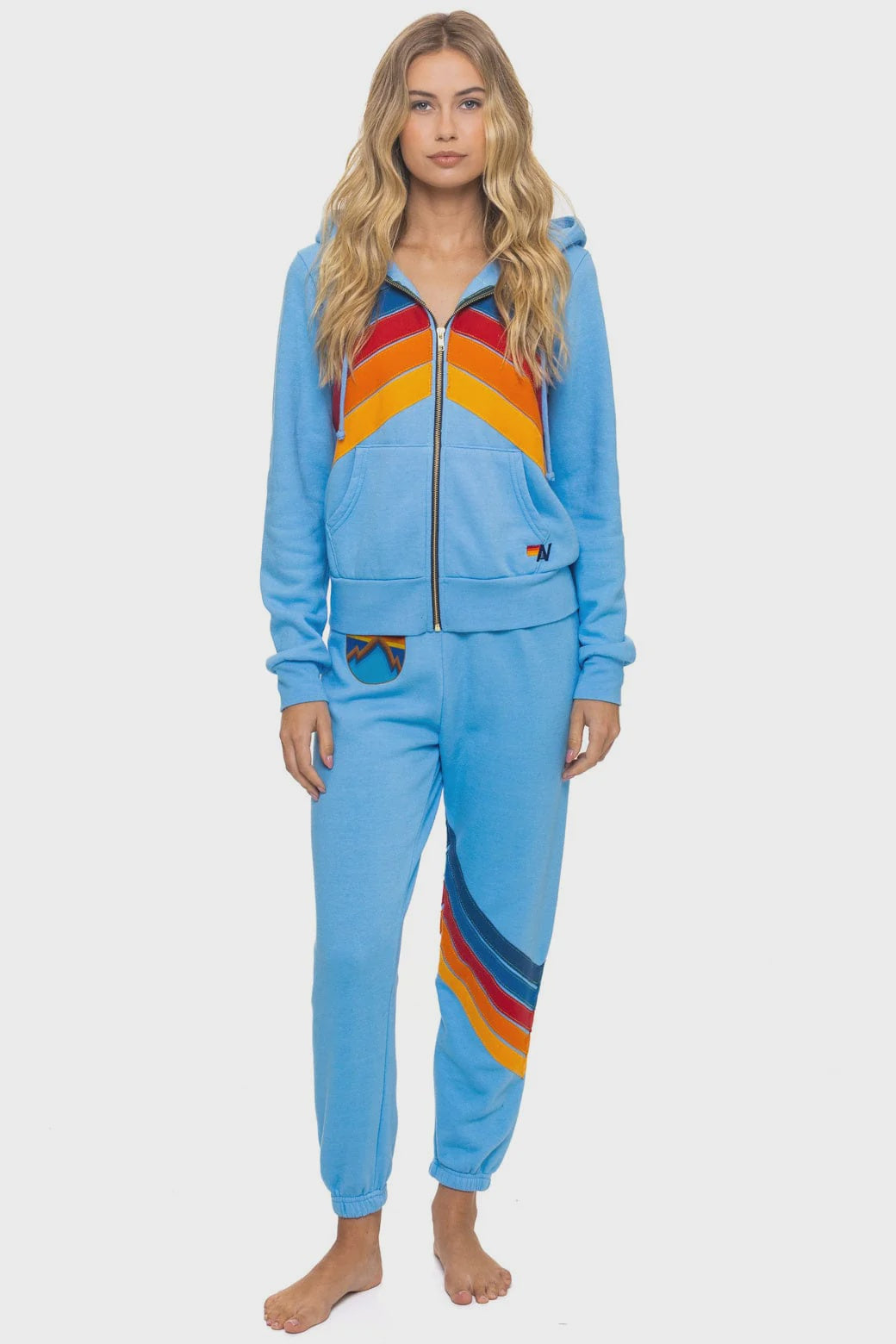 A woman with long wavy blonde hair models Aviator Nation’s all seasons circle chevron women’s sweatpant, featuring bold orange, red, and navy stripes. She wears a matching light blue zip-up hoodie and stands barefoot against a plain white background.