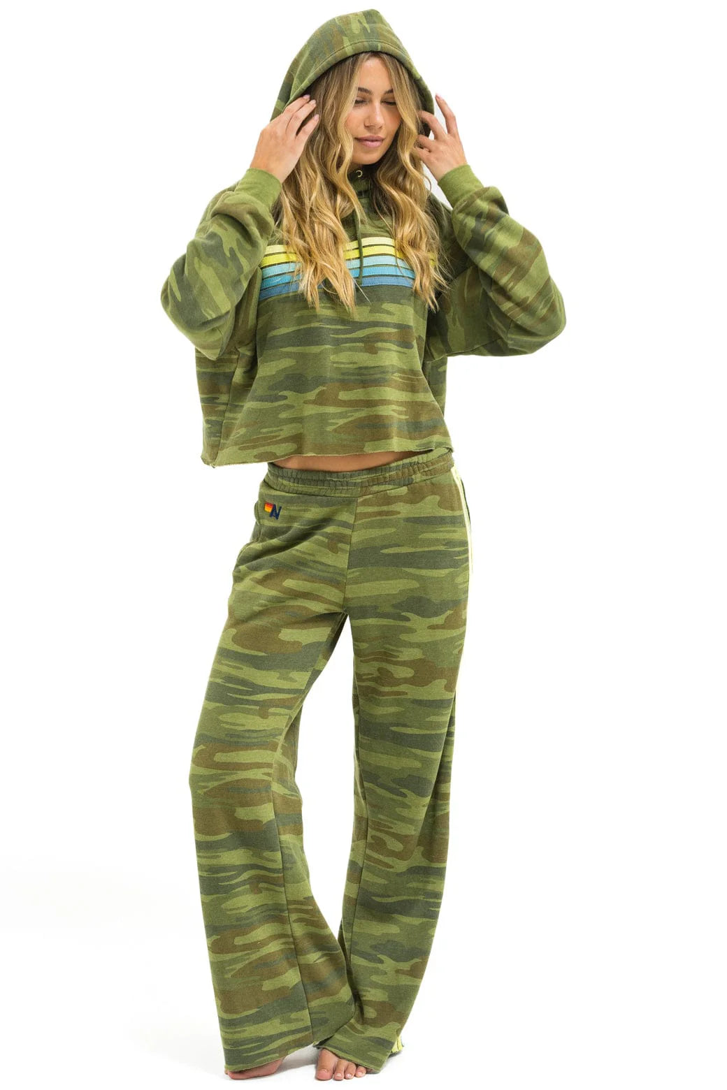 A woman with long blonde hair poses barefoot against a white background, wearing the Aviator Nation 5 stripe cropped pullover hoodie relaxed in green camouflage with blue and yellow stripes, paired with matching pants and holding the hood over her head.
