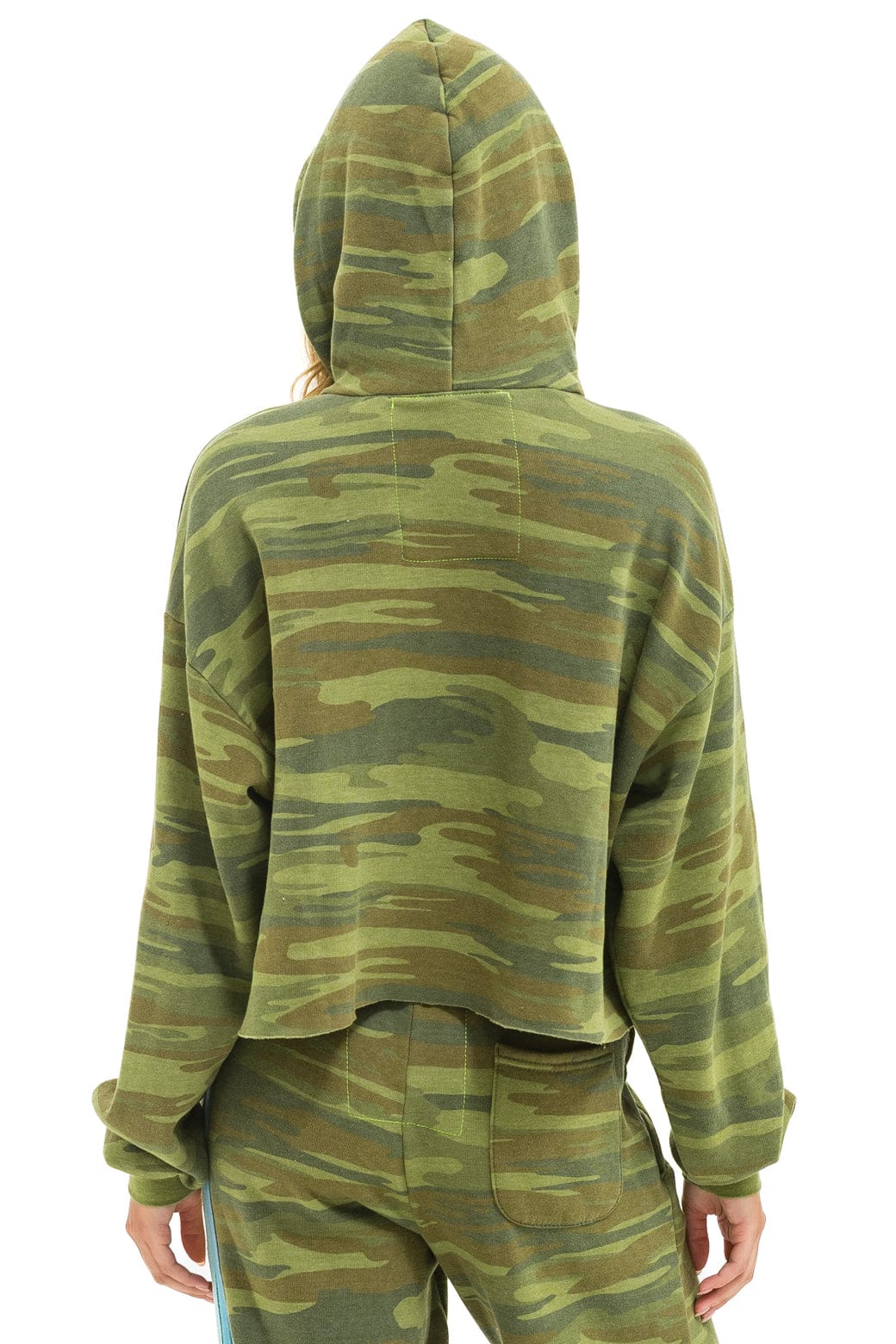 Seen from behind, a person wears the Aviator Nation 5 Stripe Cropped Pullover Hoodie Relaxed in a comfortable camouflage vintage style, hood up and arms relaxed at their sides with matching pants.