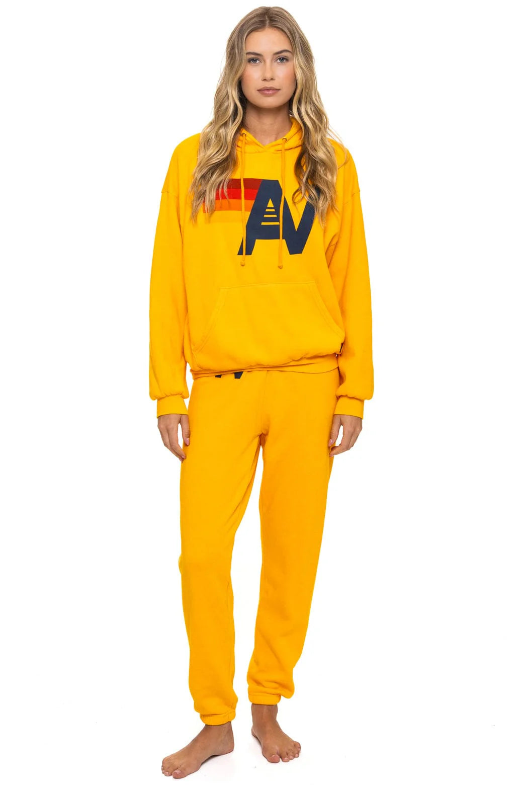 A woman with long blonde hair wears the Aviator Nation logo - pullover hoodie relaxed in bright yellow, featuring a bold "AV" logo with red and orange stripes, paired with matching sweatpants. She is barefoot against a white background.