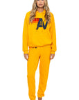 A woman with long blonde hair wears the Aviator Nation logo - pullover hoodie relaxed in bright yellow, featuring a bold "AV" logo with red and orange stripes, paired with matching sweatpants. She is barefoot against a white background.