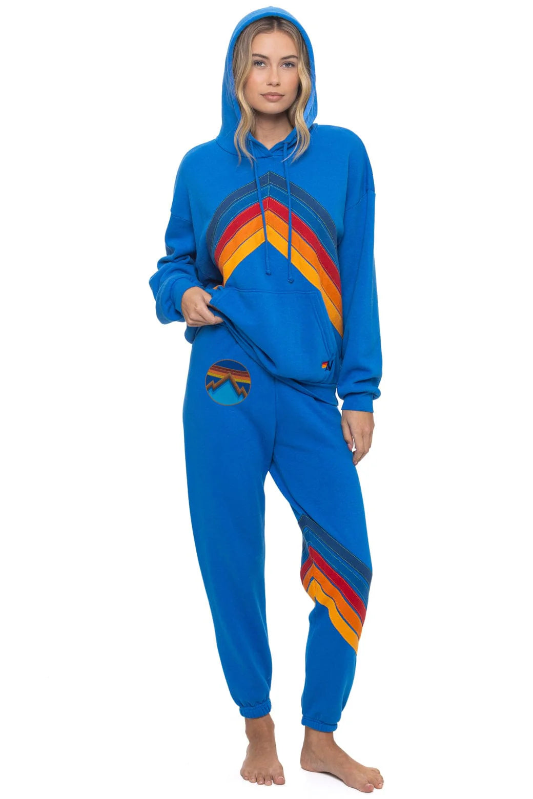 A woman stands barefoot in Aviator Nation's all seasons circle chevron women's sweatpant, a comfy vintage-inspired set with colorful rainbow stripes and a matching patch on the waistband. One hand is in her pocket, the other holding her hoodie.